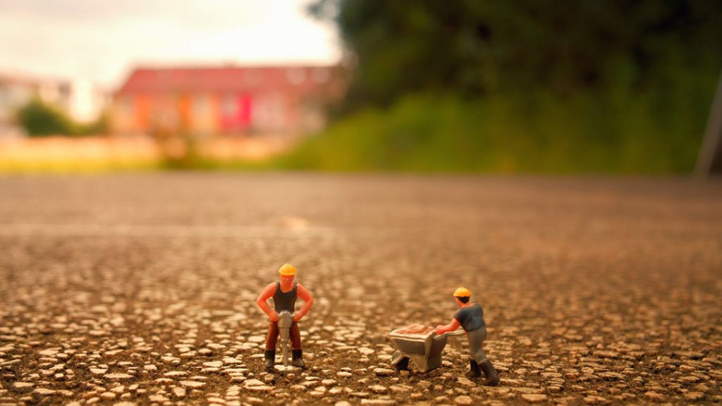 Two tiny figurines of construction workers stand on an asphalt road